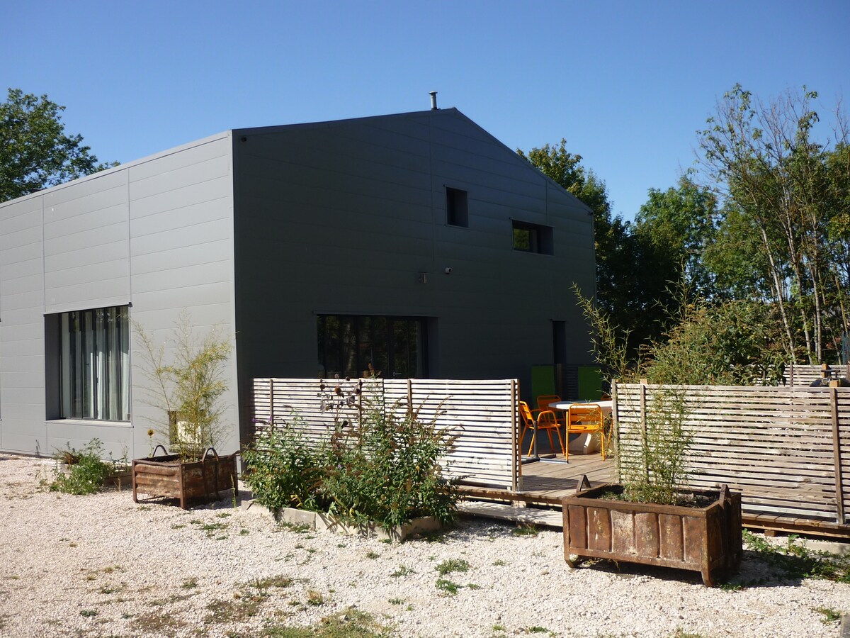 The exterior of the property showcases a modern design with a grey façade and large windows. A wooden fence separates a small outdoor seating area featuring colorful chairs, enhancing the inviting outdoor space surrounded by greenery and gravel.