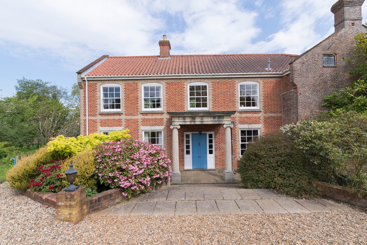 A charming Georgian farmhouse is presented, featuring a symmetrical facade with large windows and a welcoming blue front door. Lush greenery and colorful flower beds frame the entrance, and a gravel path leads to the front door, inviting guests to explore further.