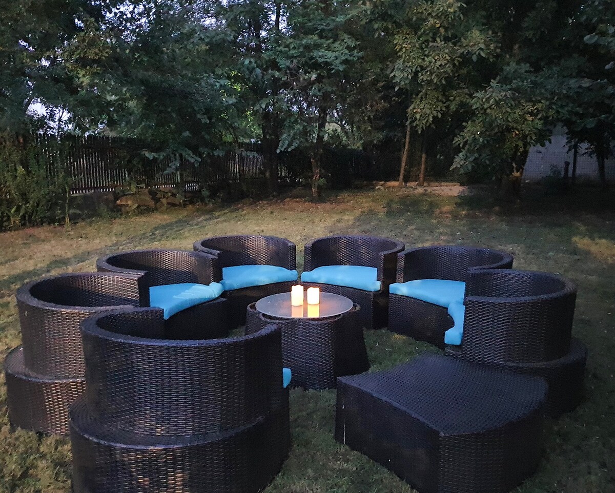 An outdoor seating area features a circular arrangement of black wicker chairs, each adorned with soft blue cushions. A small table is situated in the center, holding three lit candles, creating a warm ambiance amidst greenery and trees in the background.