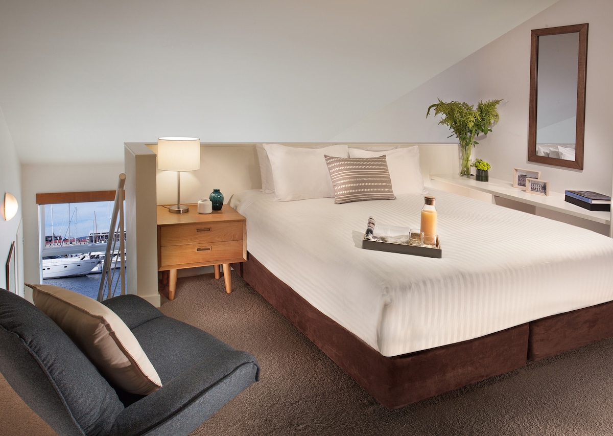 A loft-style bedroom features a spacious bed with crisp white linens and decorative pillows. A wooden bedside table holds a lamp and a small vase with greenery. A cozy chair sits nearby, and natural light streams in from a sloped window showcasing views of the marina.