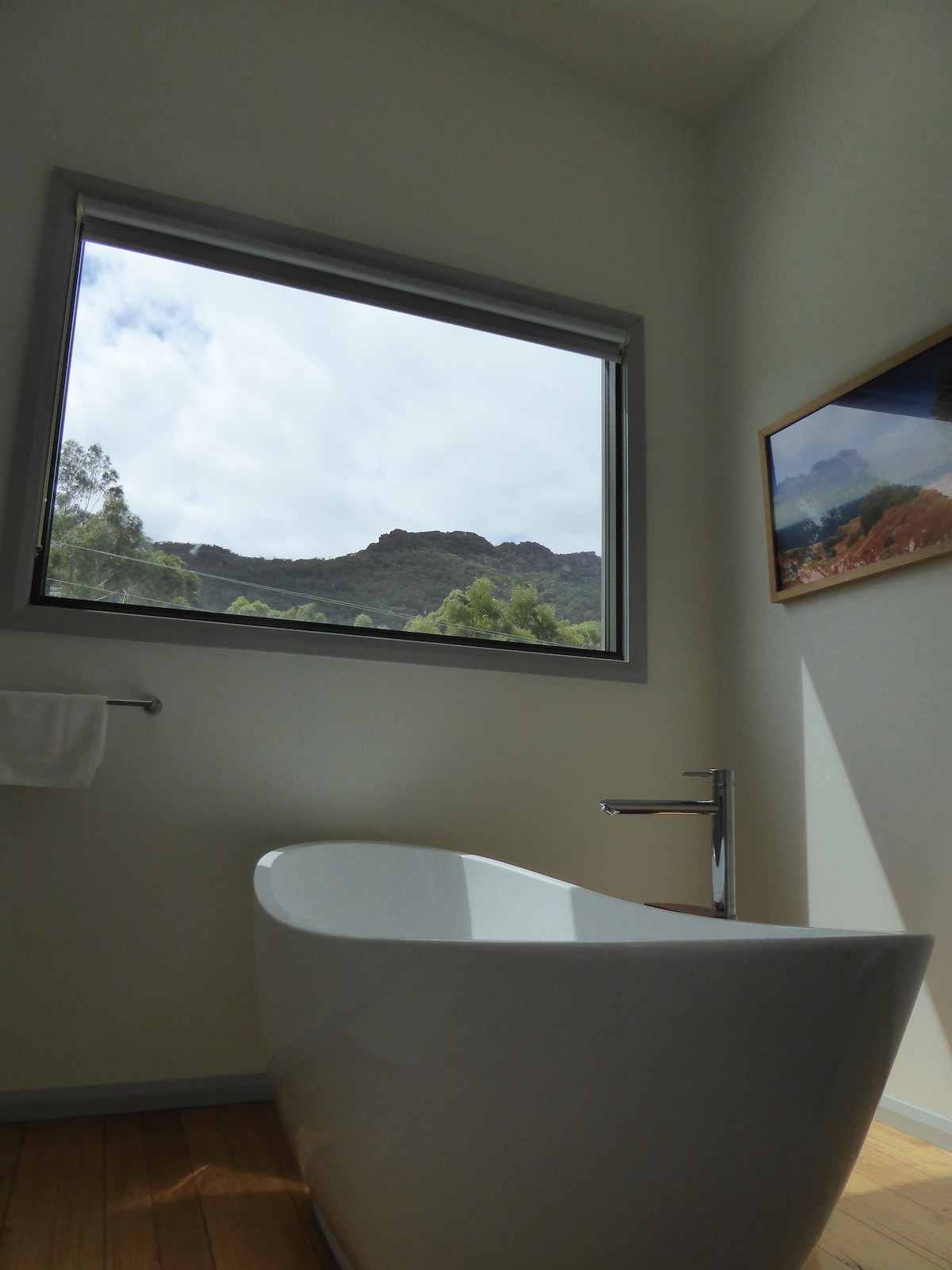 A modern bathtub is positioned in front of a large window, offering views of the surrounding mountains. Soft natural light enters the space, highlighting the clean lines of the tub and the minimalistic decor. A framed photograph hangs on the wall, complementing the serene atmosphere.