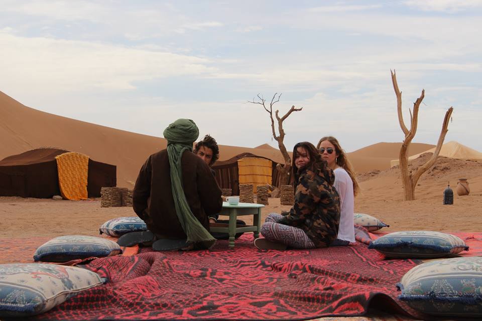 A low wooden table is surrounded by decorative floor cushions on a patterned rug. In the background, nomadic tents are visible against the sandy dunes and scattered trees. The scene captures a relaxed gathering in a serene desert landscape.
