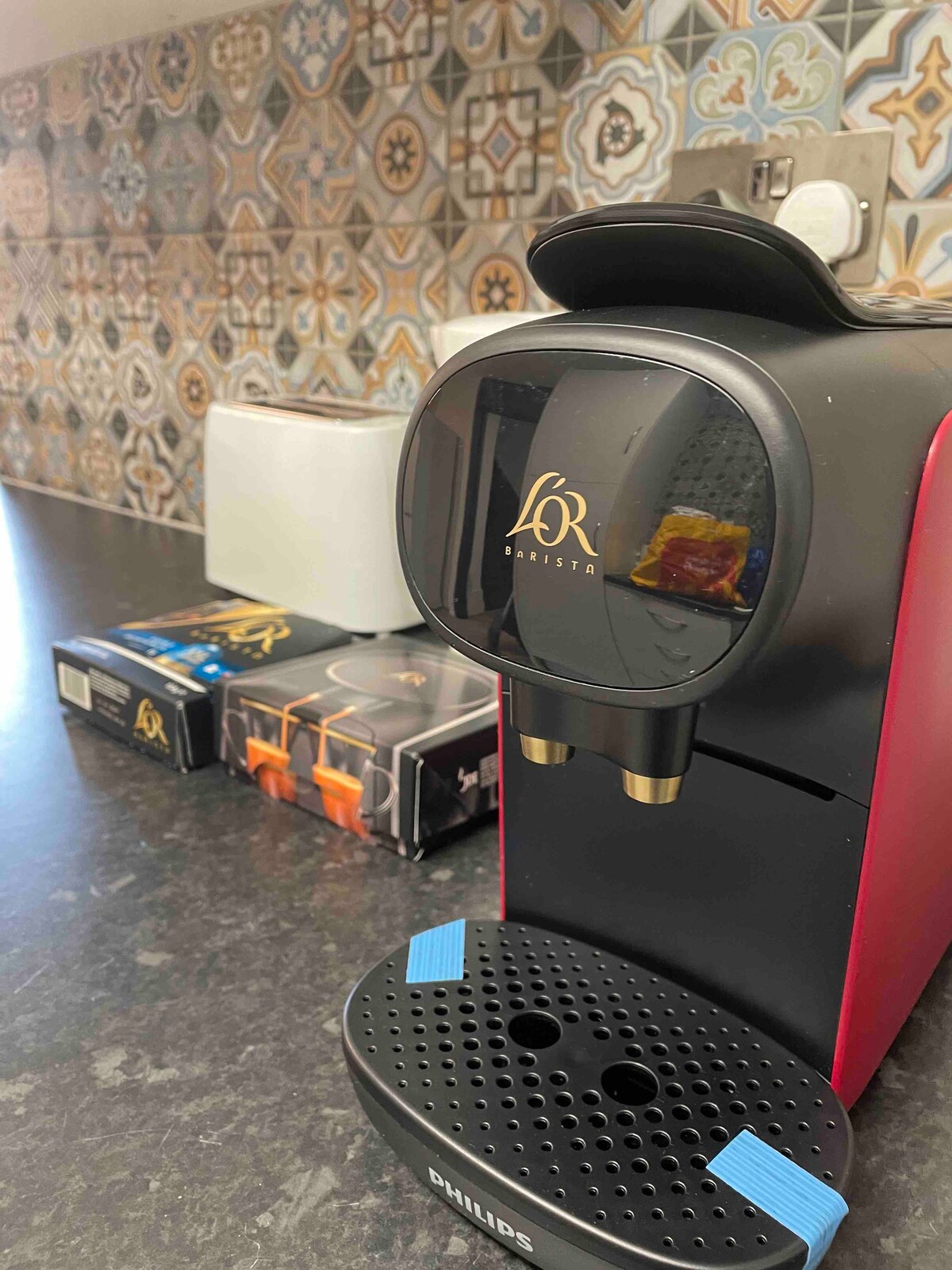 A modern coffee machine is positioned on a dark countertop. Behind it, decorative tiles with intricate patterns offer a stylish backdrop. Nearby, a white toaster and a box containing coffee pods are visible, adding convenience to the kitchen space.