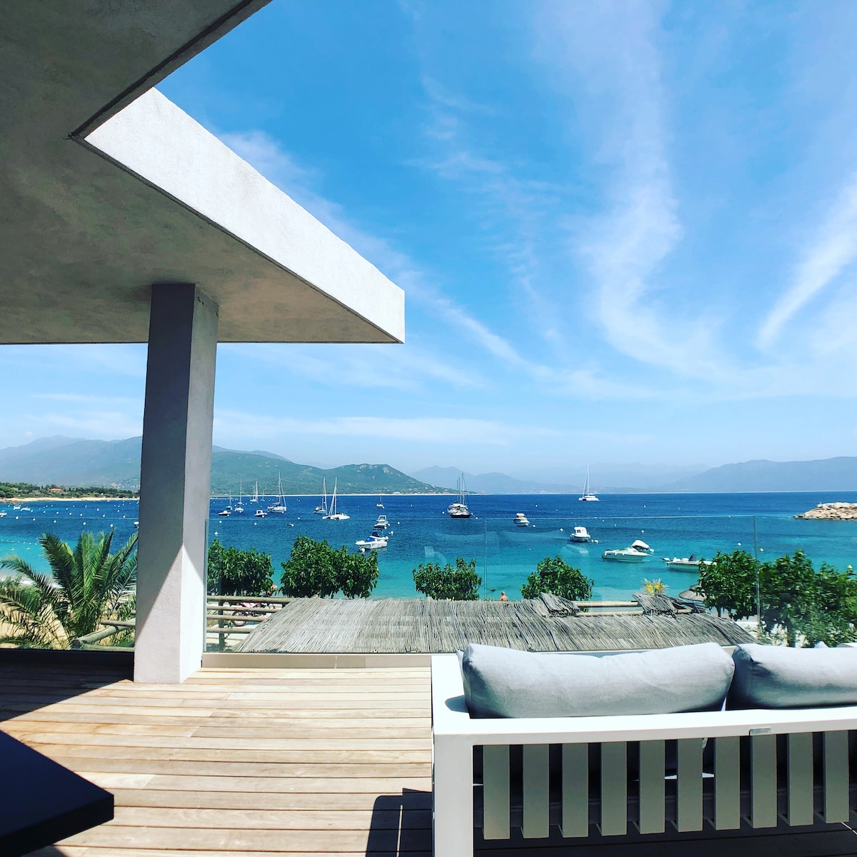 A spacious terrace offers a panoramic view of the azure sea, dotted with sailboats and framed by distant mountains. Comfortable outdoor furniture is arranged for relaxation, under a bright blue sky with scattered clouds, enhancing the connection to the serene coastal environment.