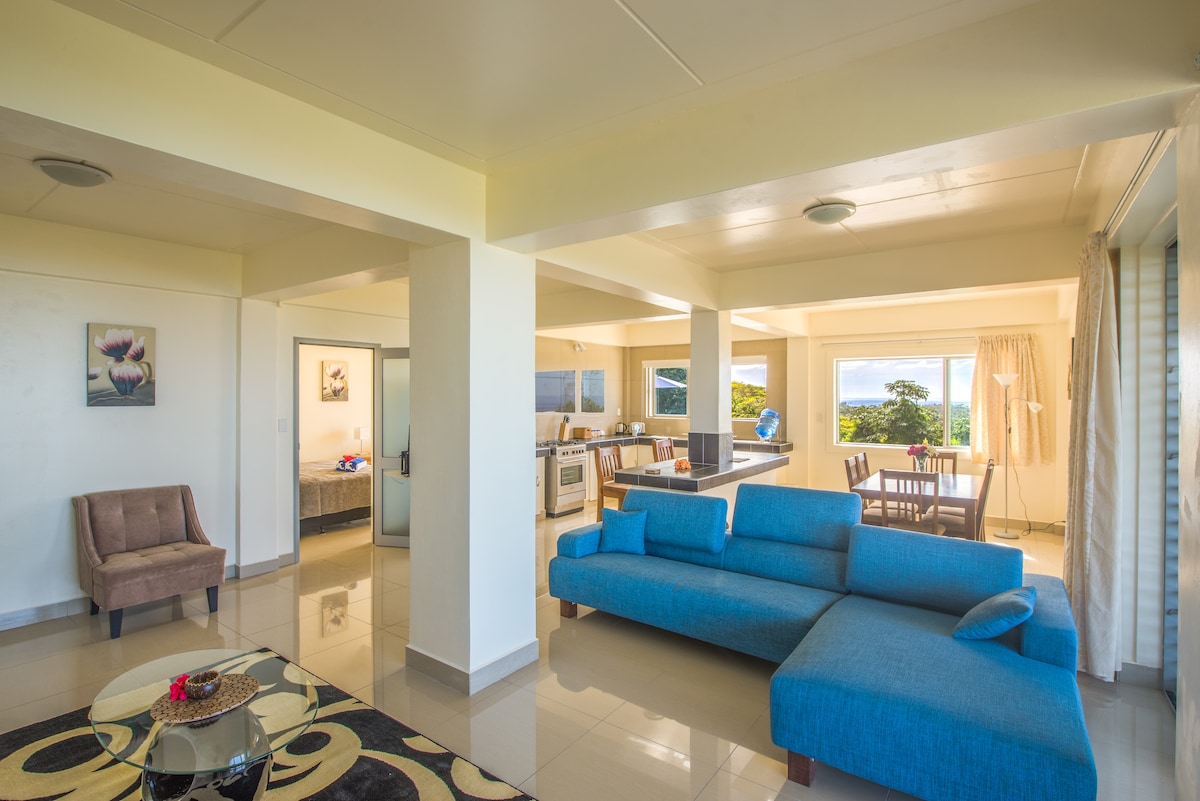 An open-plan living area features a blue sectional sofa and modern glass coffee table. Natural light floods the space from large windows, highlighting the kitchen and dining area in the background. Artwork adorns the walls, complementing the neutral color scheme.