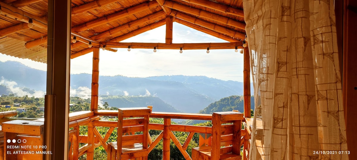A private balcony is framed by bamboo beams, offering stunning views of misty mountains in the distance. Wooden chairs are positioned for relaxation, and soft morning light spills into the space, creating a serene atmosphere. Decorative string lights enhance the cozy setting.