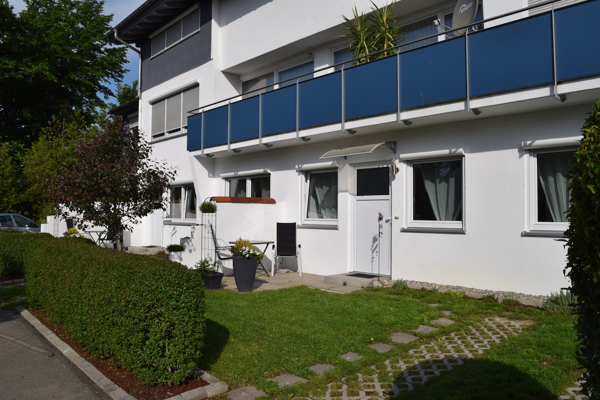 A modern building exterior features a white facade complemented by blue balcony railings. A small terrace with potted plants and a pathway lined with greenery is visible in front, providing an inviting entrance to the property.