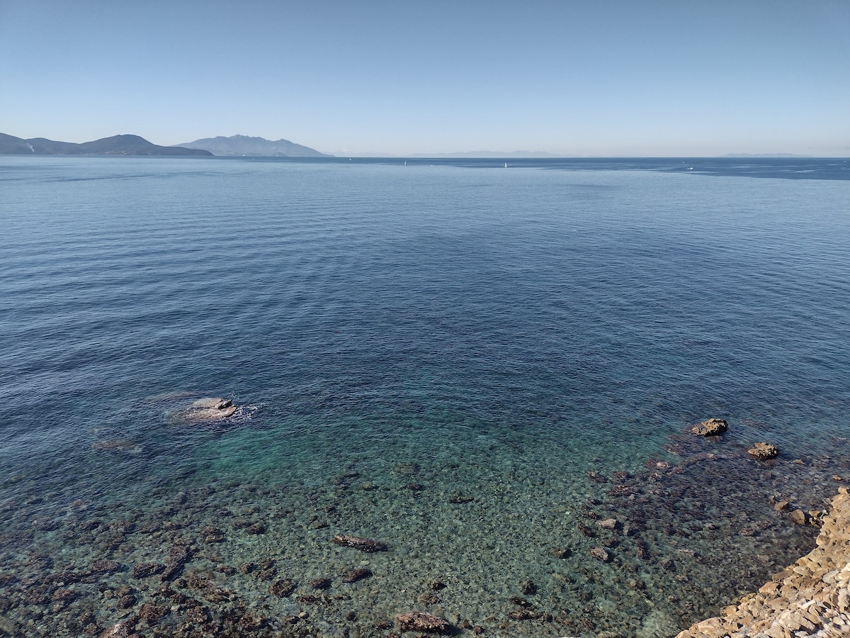 A calm seascape is presented, showcasing clear blue waters transitioning to rocky sea beds. The horizon meets distant islands under a clear sky, with gentle waves creating a soothing ripple effect on the surface. The tranquil environment highlights the natural beauty of the coastline.