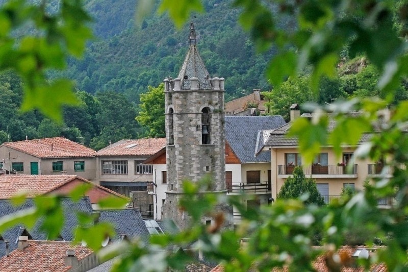 A historic stone tower rises amidst a cluster of buildings with varying roof styles. Surrounding trees and foliage frame the view, creating a natural border that highlights the architecture of the village nestled in the mountains.