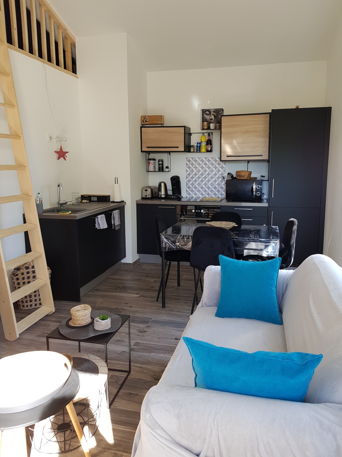 The open kitchen area is equipped with modern appliances and cabinetry, featuring a glass dining table surrounded by four chairs. A cozy seating arrangement with a light-colored sofa and colorful cushions is positioned nearby, harmonizing with the wood-toned flooring and bright wall accents.