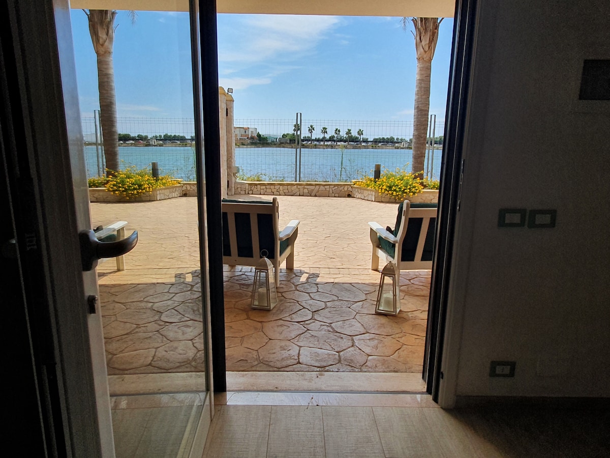 A view of the tranquil outdoor area is visible through sliding glass doors. Two lounge chairs are positioned on a patio, framed by palm trees and bordered by colorful flower beds. A peaceful body of water reflects the surrounding landscape.