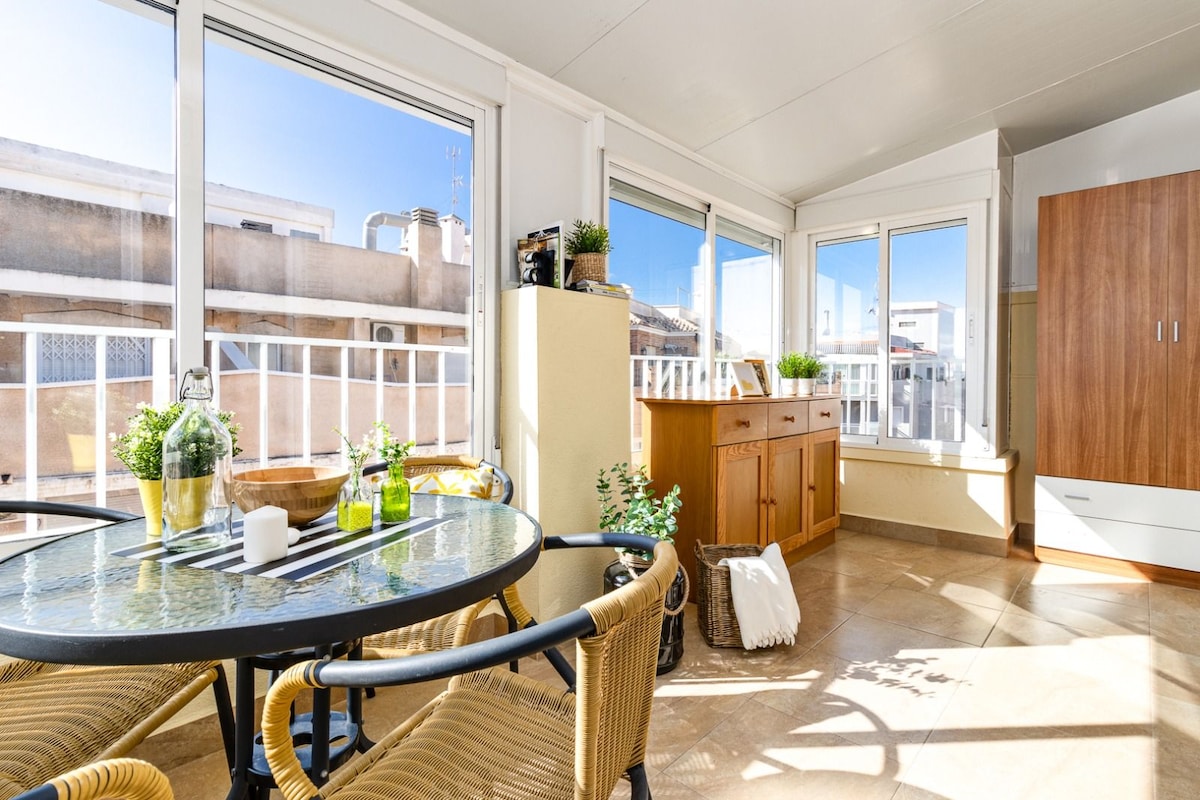 A bright corner of the terrace is shown, featuring a round glass table surrounded by woven chairs. Large windows allow natural light to flood the space, while potted plants add a touch of greenery to the area. Wooden cabinets are visible in the background.