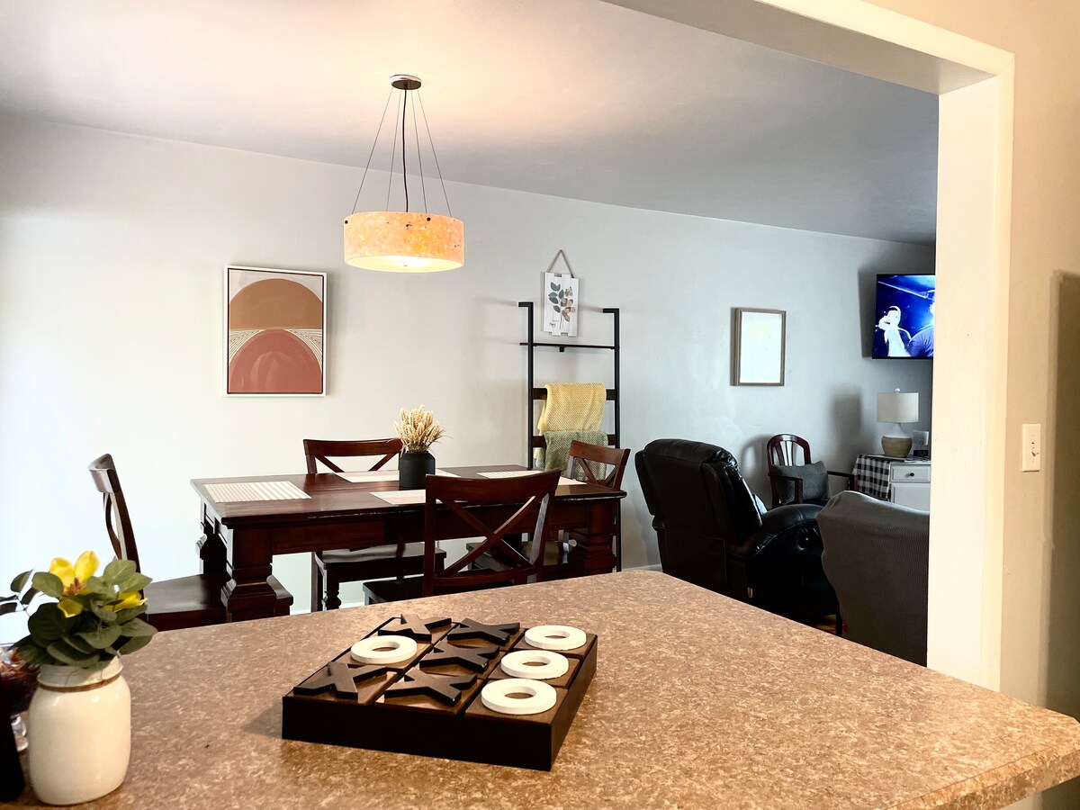 A spacious dining area is visible, featuring a polished wooden table with chairs. A light fixture hangs overhead, and a TV is mounted on the wall. Adjacent seating includes a cozy couch and armchair, creating an inviting gathering space.