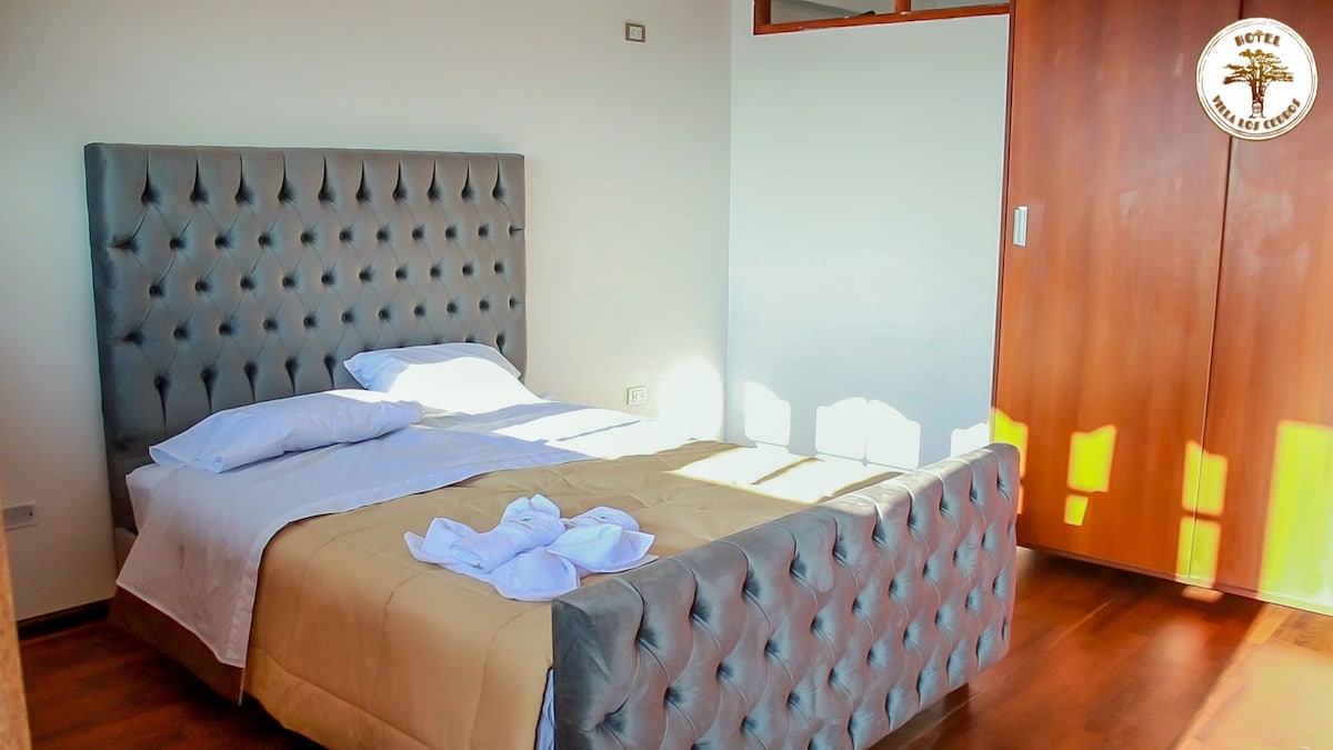 A cozy bedroom features a tufted gray bed frame with a light-colored blanket draped over a spacious bed. Natural light filters through a nearby window, casting patterns on the wooden floor. A wooden closet is visible, providing additional storage.