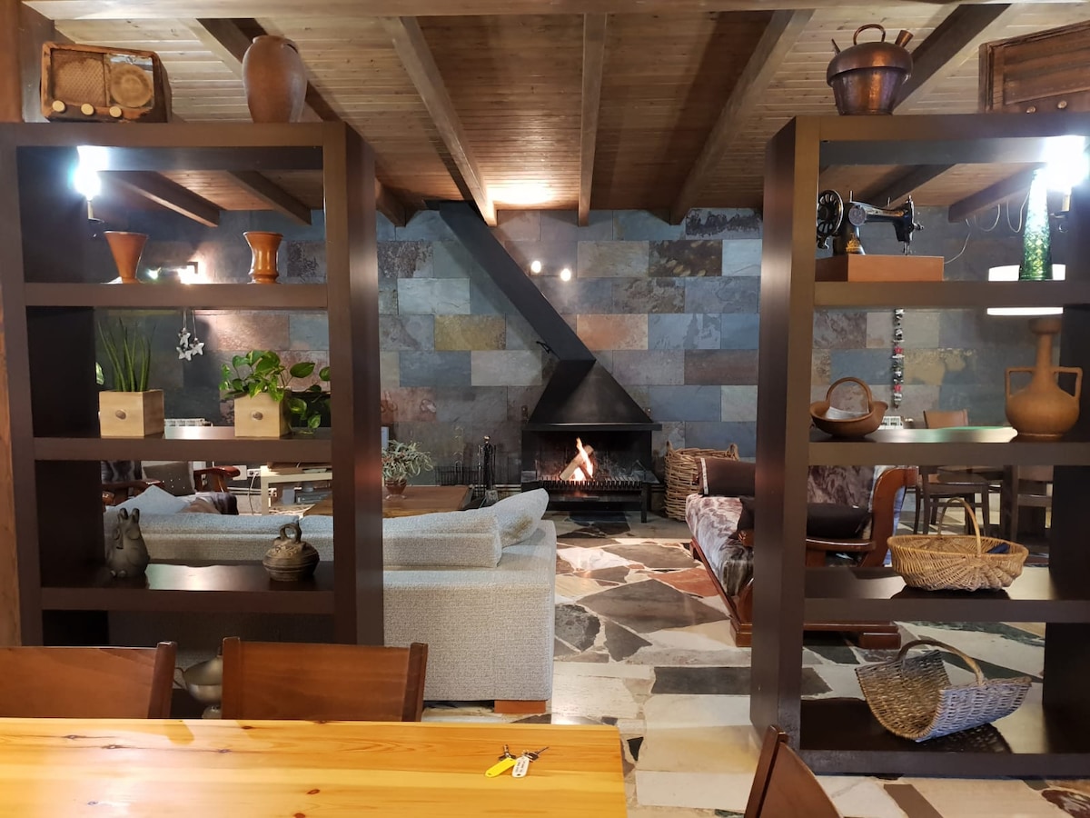 A spacious living area is defined by wooden beams overhead and a stone fireplace, providing a cozy focal point. Bookshelves are arranged with decorative items, while comfortable seating invites relaxation. Natural textures and colors are present throughout, enhancing the overall aesthetic of the space.