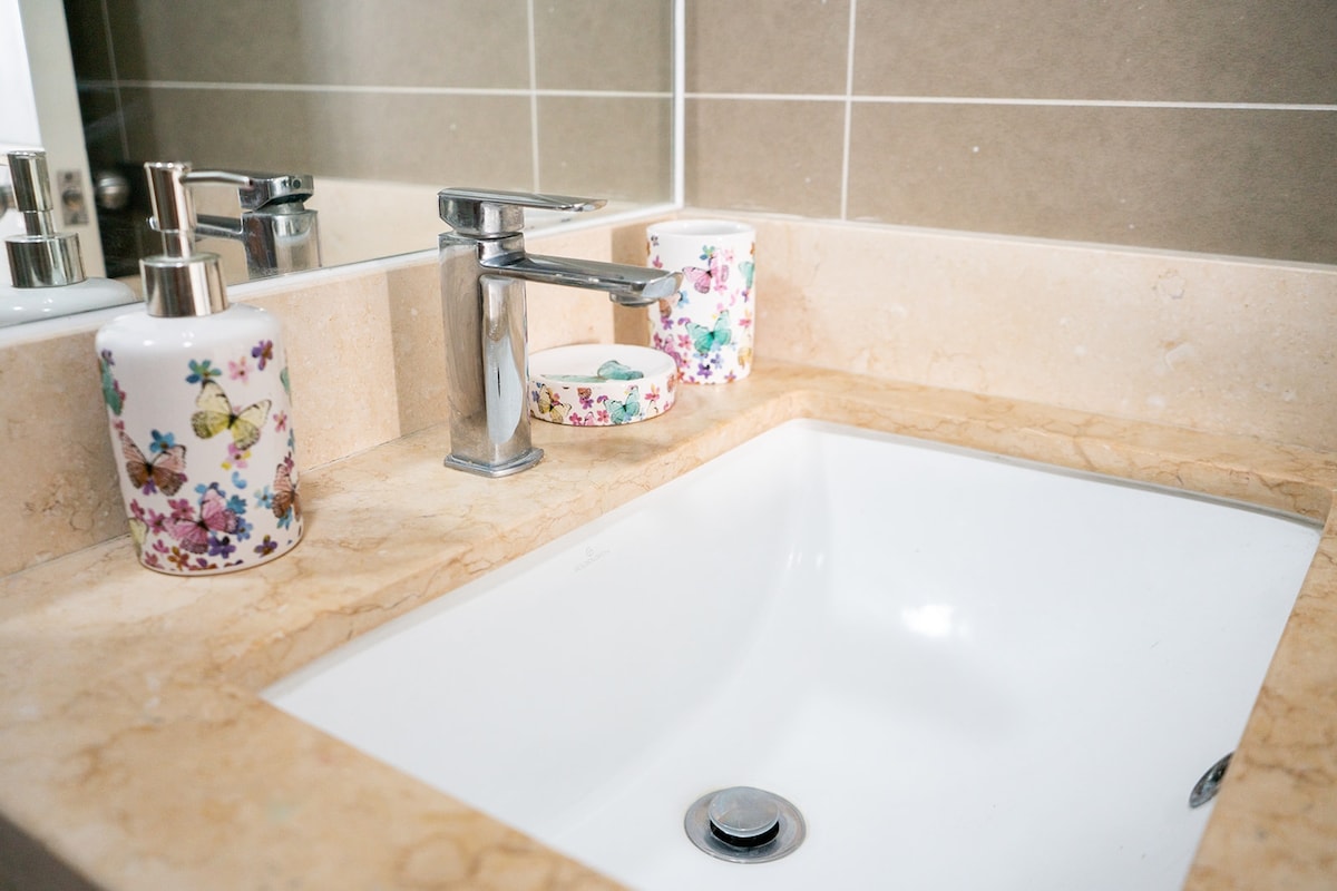 A clean and modern bathroom sink area features a marble countertop with a white basin. Decorative soap dispensers with floral designs enhance the space. Wall tiles in neutral tones create a calming backdrop, and a large mirror above the sink reflects the contemporary fixtures.