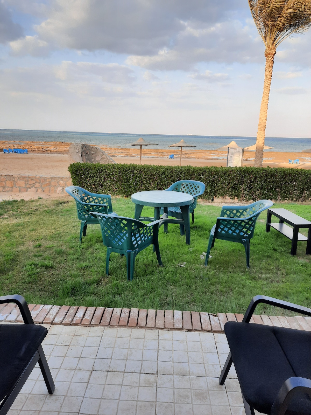 A outdoor seating area is featured, consisting of a round table surrounded by four green chairs on a grassy patch. In the background, a sandy beach is visible along with umbrellas and palm trees, providing a coastal view.