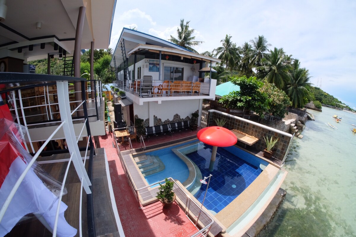 A view from above showcases a pool area with a red mushroom-shaped feature, surrounded by seating options. Several lounge chairs line the edge, and tropical foliage adds greenery. The shoreline is visible, offering a sense of proximity to the water.