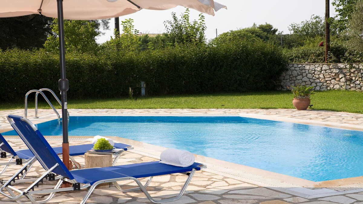 A tranquil pool area is highlighted by a clear blue pool, framed by lush greenery. Two lounge chairs with sun umbrellas are positioned nearby, accompanied by a small side table and decorative plants. The stone patio provides ample space for relaxation and enjoyment.