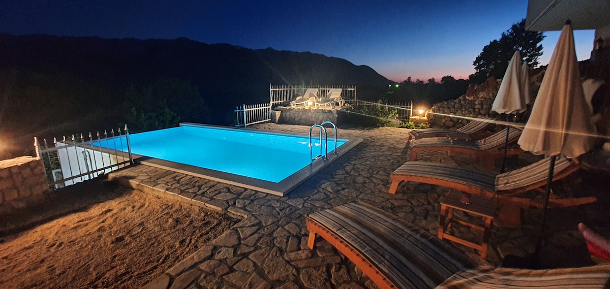 A saltwater pool is illuminated in soft blue hues under a twilight sky, surrounded by lounge chairs and umbrellas. The serene outdoor space offers views of the mountains, creating a peaceful retreat for relaxation.