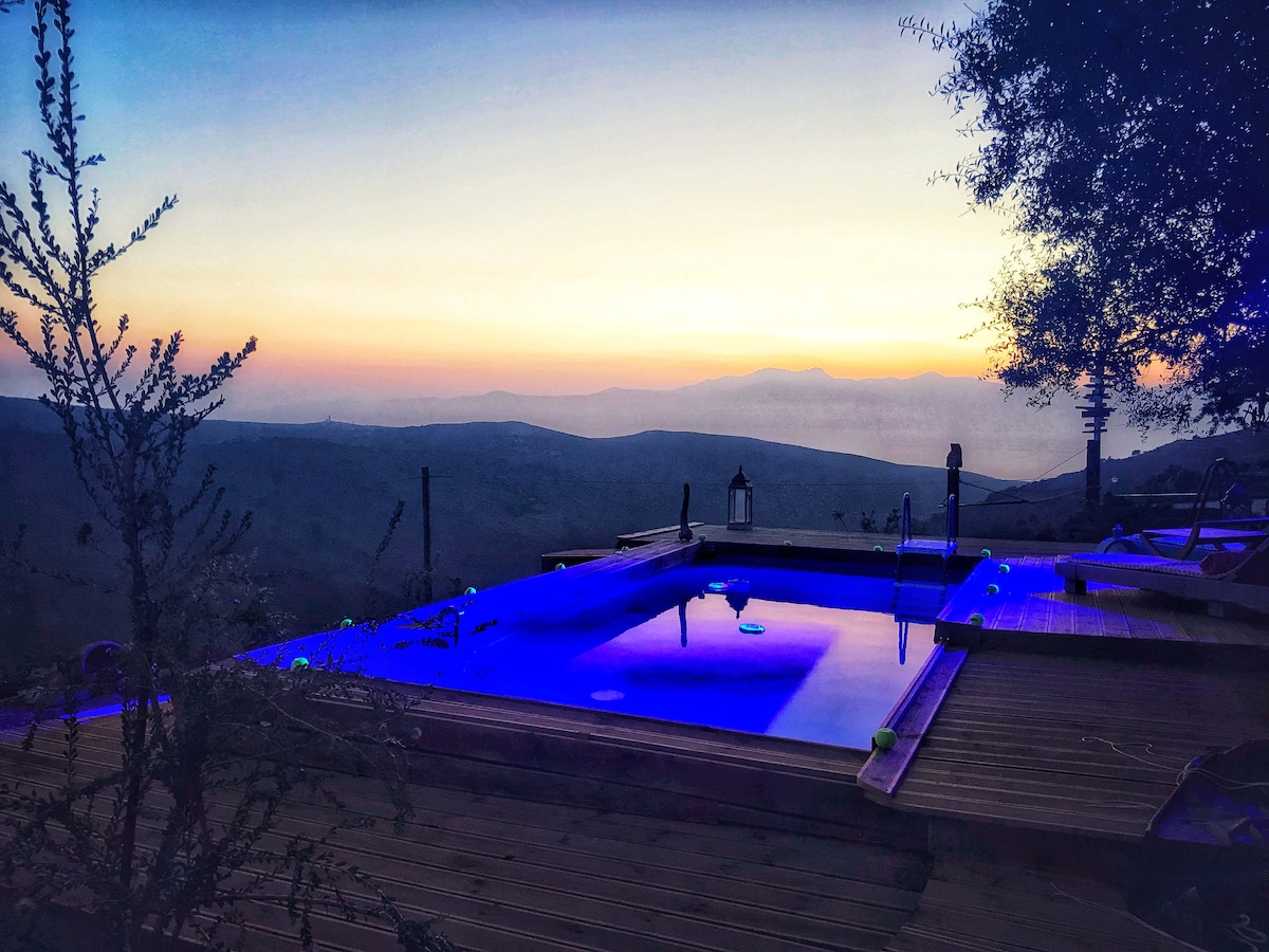 A serene outdoor area presents a swimming pool, illuminated with soft blue lighting, set against a backdrop of a sunset sky. Rolling hills are visible in the distance, while the surrounding nature adds a tranquil touch to the scene.