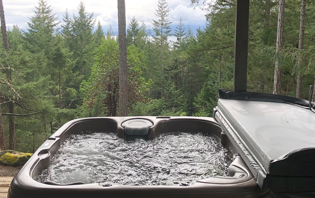 A private hot tub is situated on a wooden deck, surrounded by lush greenery and tall trees. The water is bubbling gently, with views of the ocean visible through the foliage in the background.