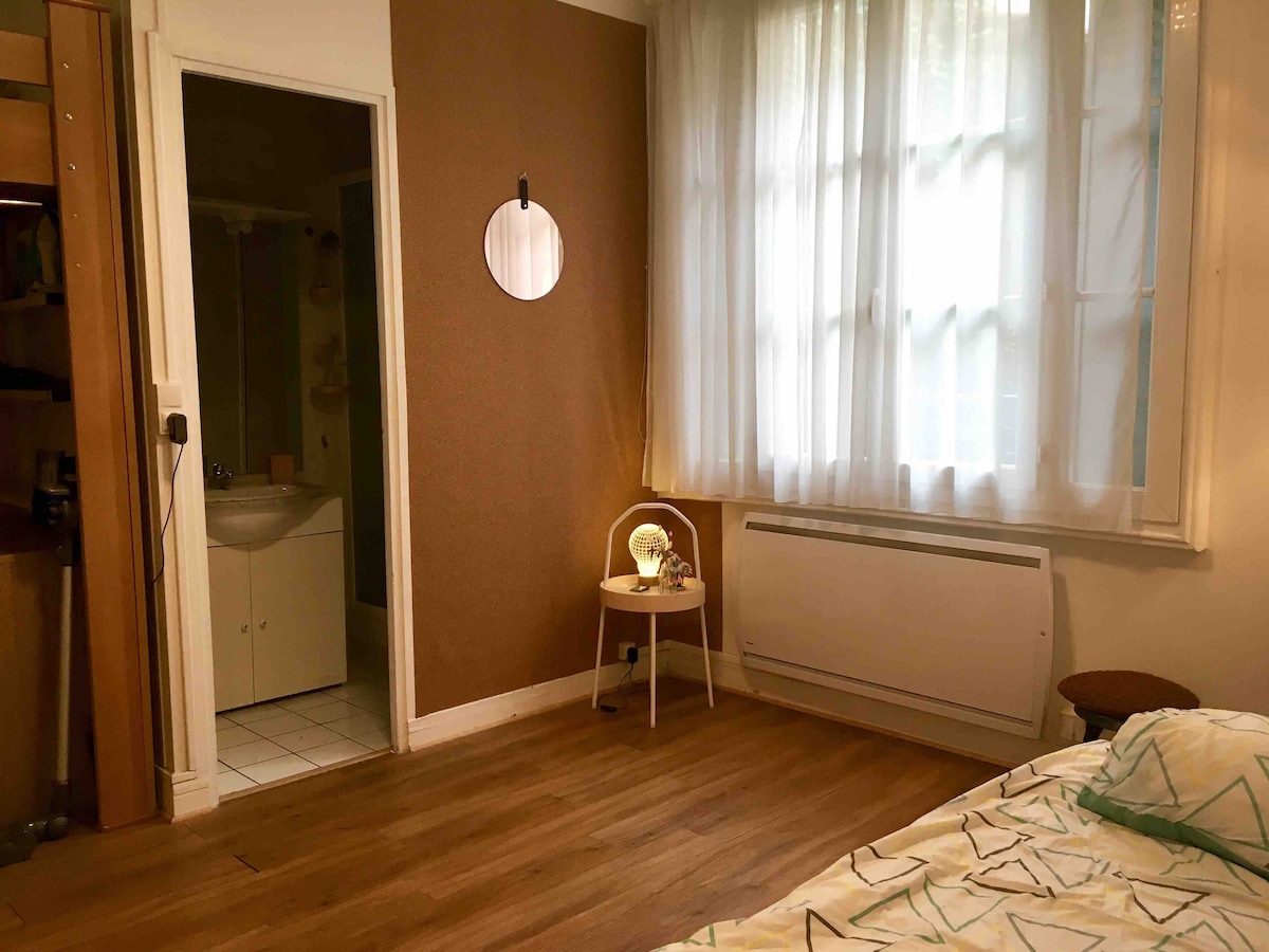 A cozy studio space is showcased, featuring warm wood flooring and natural light filtering through sheer curtains. A mirror is displayed on the wall, while a small table with a lamp rests beside a comfortable chair, creating a charming corner.