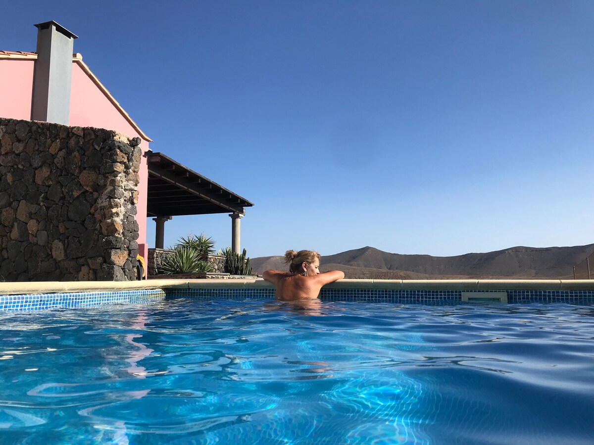 A private pool with clear water reflects the surrounding landscape. The structure features a rustic stone wall and a shaded terrace, providing a serene escape. Mountains can be seen in the distance, framed by a clear blue sky.