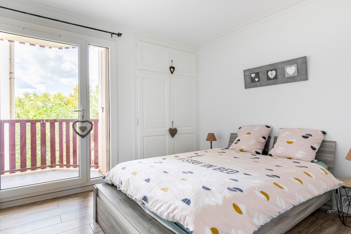 A spacious bedroom is presented, featuring a large bed with a patterned duvet cover. Two bedside lamps are positioned on either side of the bed. A sliding glass door leads to a balcony, allowing natural light to fill the room while offering views of the greenery outside.