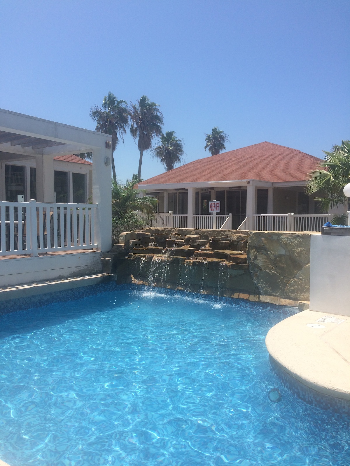 A sparkling pool is featured with a rock waterfall, providing a relaxing ambiance. Surrounding palm trees add a tropical feel, while a covered pavilion with a red roof is visible in the background, enhancing the outdoor space's inviting nature.