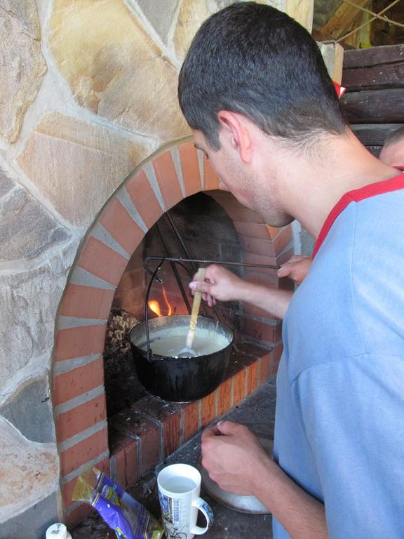 A person is stirring a pot hanging over a fire in a rustic fireplace. The warm glow of the flames reflects off the surrounding stone structure. A white mug is placed on the surface nearby, adding a homey touch to the setting.