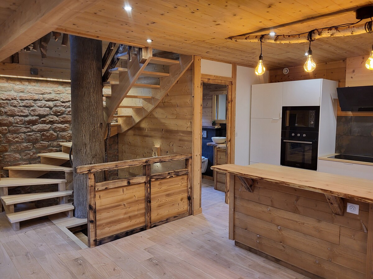 A modern kitchen is complemented by wooden cabinetry and a central island. A spiral staircase ascends to the upper level, flanked by a rustic stone wall. Pendant lighting adds illumination, enhancing the warm wooden interior, which embodies a cozy and functional living space.