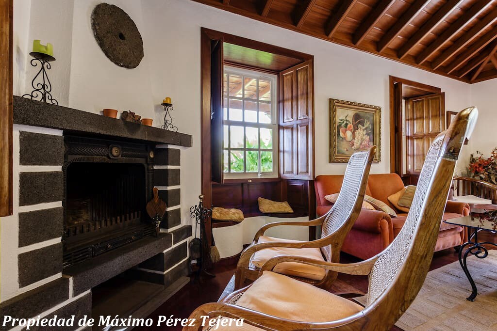 A cozy living room features a rustic stone fireplace as the focal point. Two comfortable chairs sit nearby, surrounded by wooden framed windows that allow natural light to enter. Decorative elements like artwork and plants enhance the inviting space.
