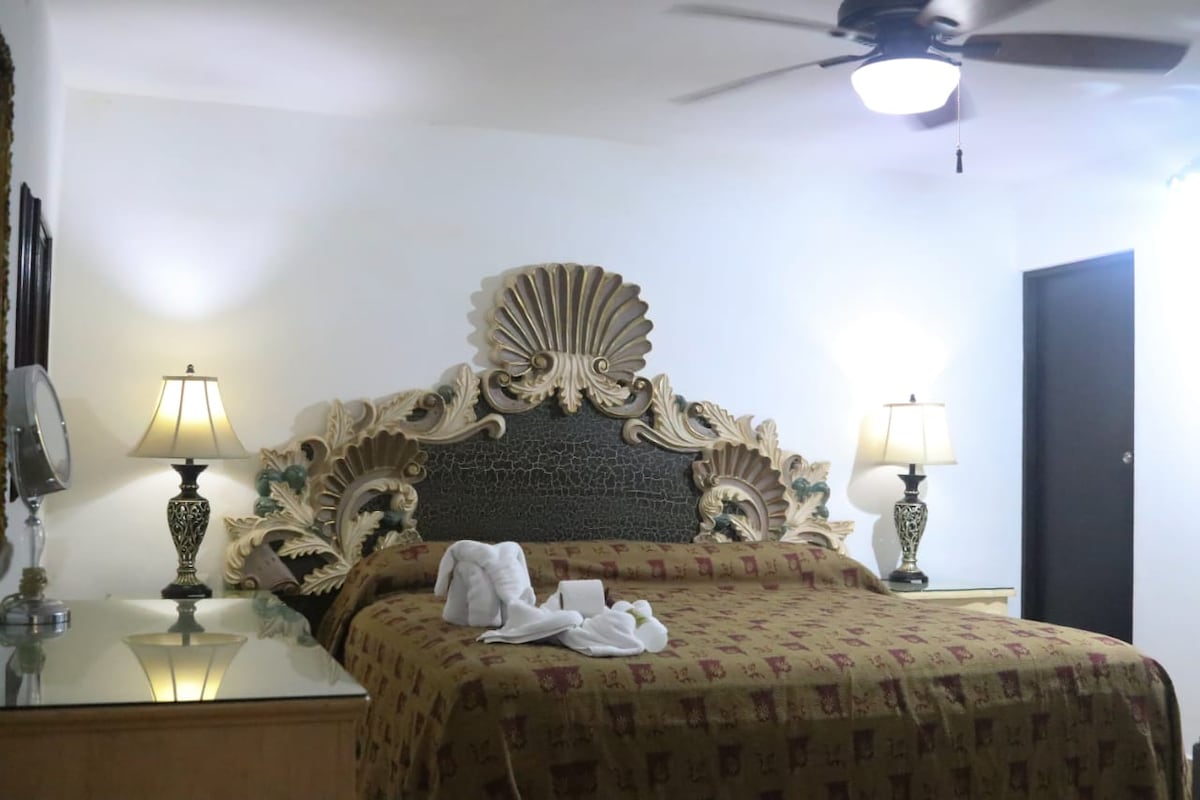 A spacious bed with an ornate headboard is complemented by decorative bedside lamps. The bedspread features intricate patterns, while light is provided by fixtures in the ceiling. A mirrored dresser adds brightness to the room, which includes a closed door in the background.