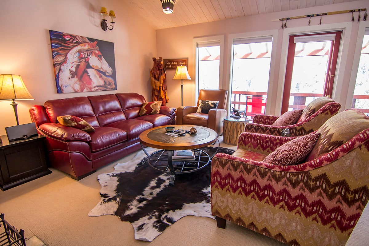 A cozy living area features a red leather sofa and armchair, along with patterned accent chairs. A round coffee table rests on a cowhide rug, with warm lighting from a nearby lamp and natural light from multiple windows.