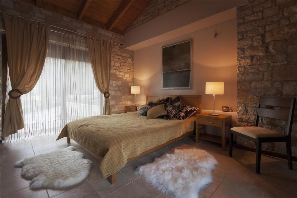 A serene bedroom features a spacious bed dressed in a brown blanket, complemented by decorative pillows. Soft lighting from bedside lamps illuminates the stone walls, while large glass doors open to the outside. Two plush rugs adorn the tiled floor, enhancing the inviting ambiance.
