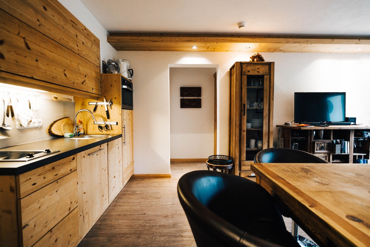 The kitchen area features warm wooden cabinetry, equipped with a sleek black countertop and essential appliances. A dining table with dark chairs is visible, and an open doorway leads to another room. Natural light highlights the wooden textures throughout the space.