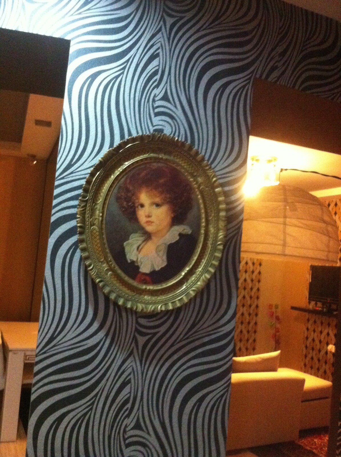 A decorative wall features bold, swirling patterns in black and white. A circular, gilded frame surrounds a classic portrait, creating a focal point. In the background, a cozy living area is visible, equipped with soft seating and a television, well-lit by gentle overhead lighting.