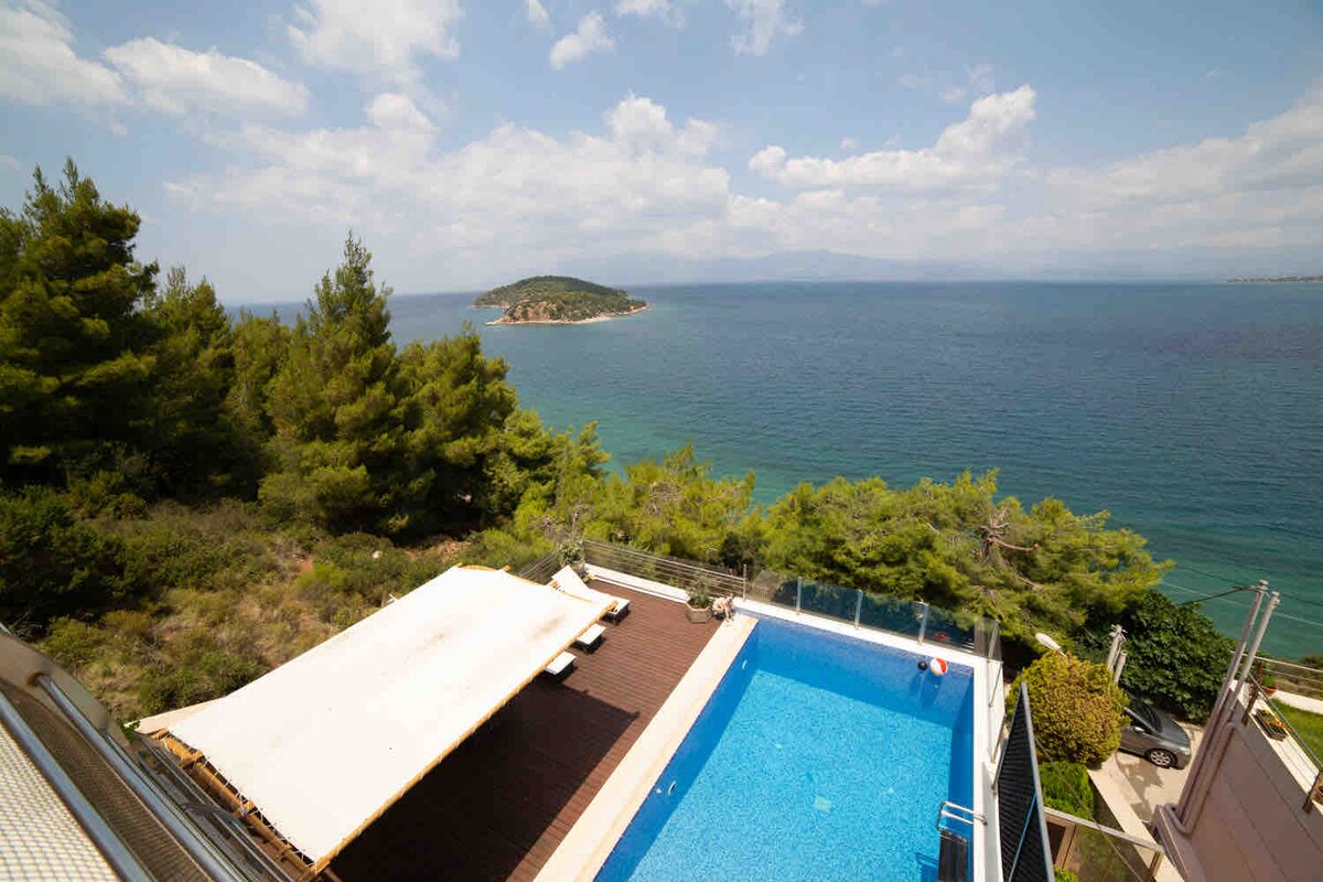 The image showcases a bright blue swimming pool overlooking the sea, with sun loungers positioned nearby under a large sunshade. Lush greenery frames the area, enhancing the natural scenery. The distant island and open sky create a serene waterfront ambiance.