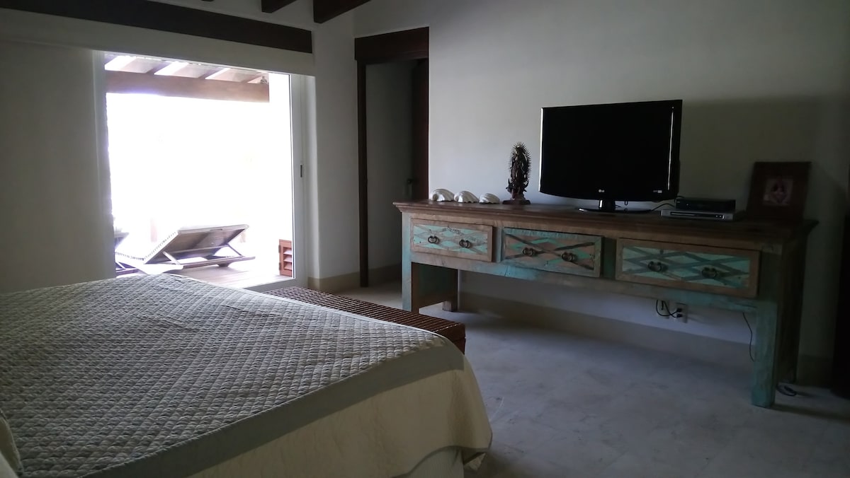 A well-furnished sleeping area features a light-colored quilted bedspread draping over a spacious bed. A wooden console table with decorative elements stands against the wall, complemented by a television. Natural light enters through large glass doors leading to an outdoor seating area.