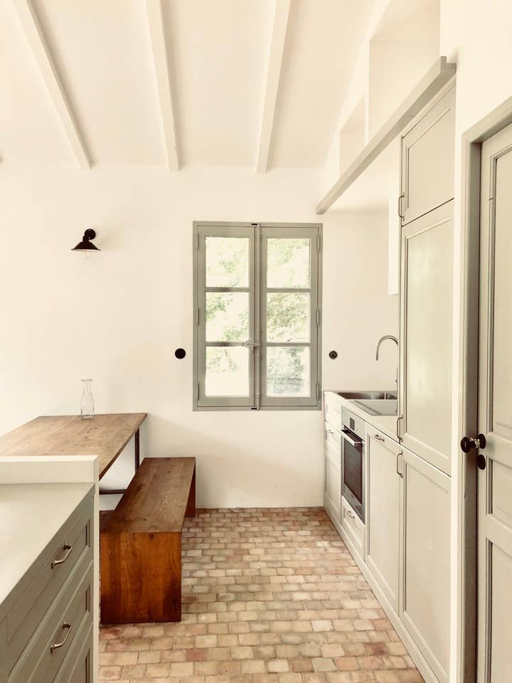 A functional kitchen space displays light-colored cabinetry and a clean layout. A wooden bench and table provide seating, while a window allows natural light to illuminate the area. The flooring features a distinctive cobblestone pattern, enhancing the rustic charm of the space.
