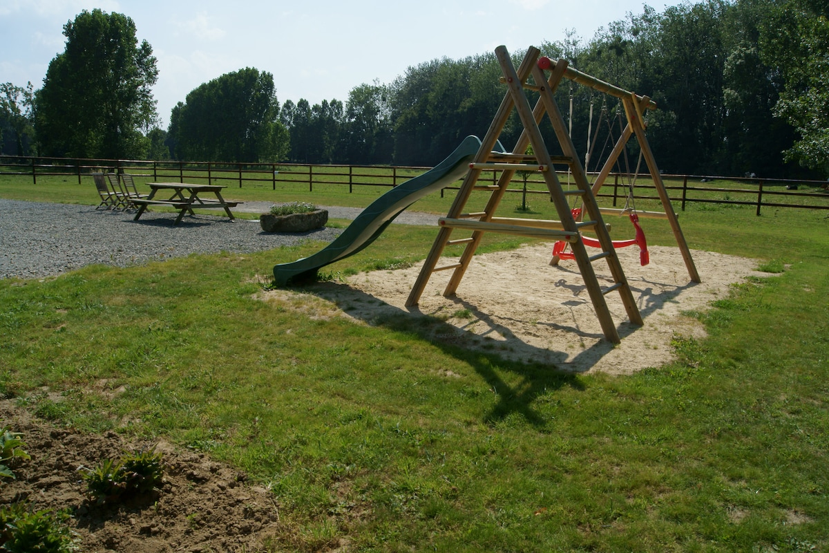 An outdoor play area is featured, complete with a wooden climbing frame, slide, and swings set in a sandy ground. Nearby, a picnic table is positioned on the grass, bordered by a gravel path and lush greenery. Fencing outlines the expansive yard.