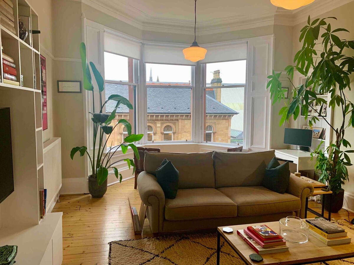 A cozy living area features a light brown sofa positioned near large bay windows. Lush green plants add a natural touch, while a light fixture hangs gracefully above. A warm wooden floor complements the contemporary bookshelf and modern decor, providing a welcoming ambiance.