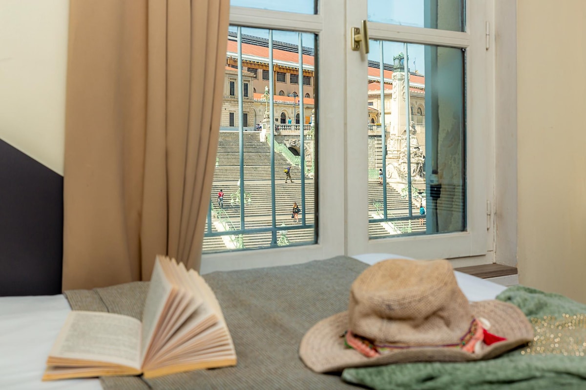A view from the room showcases the monumental staircase of Saint-Charles railway station. In the foreground, an open book and a hat rest on the spacious bed, while natural light enters through the open window, enhancing the inviting atmosphere.