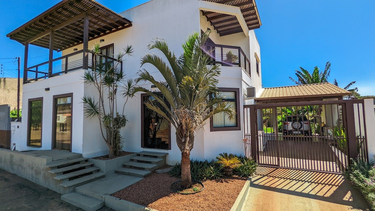A two-story house is presented with a modern design, featuring a spacious front entrance with steps leading to large windows. Palm trees and shrubs surround the building, while a covered garage with a gated entrance is visible on the side.