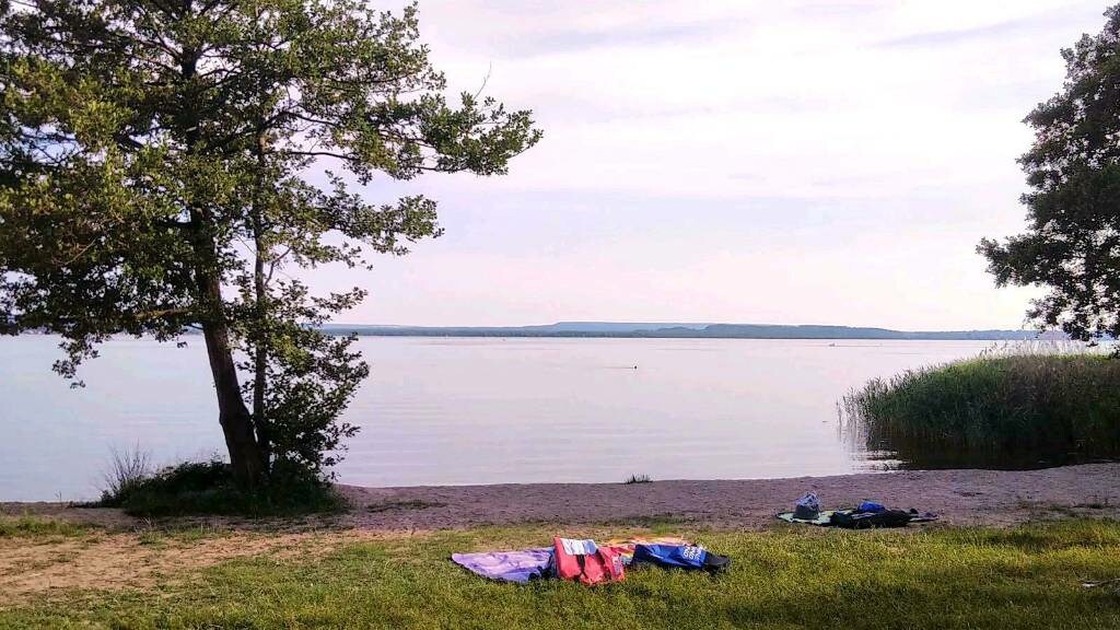 A serene lakeside scene features a smooth, sandy shore bordered by lush green grass. In the foreground, colorful towels are spread out on the sand, with gentle ripples reflecting the soft hues of the water. Trees provide partial shade along the shoreline.