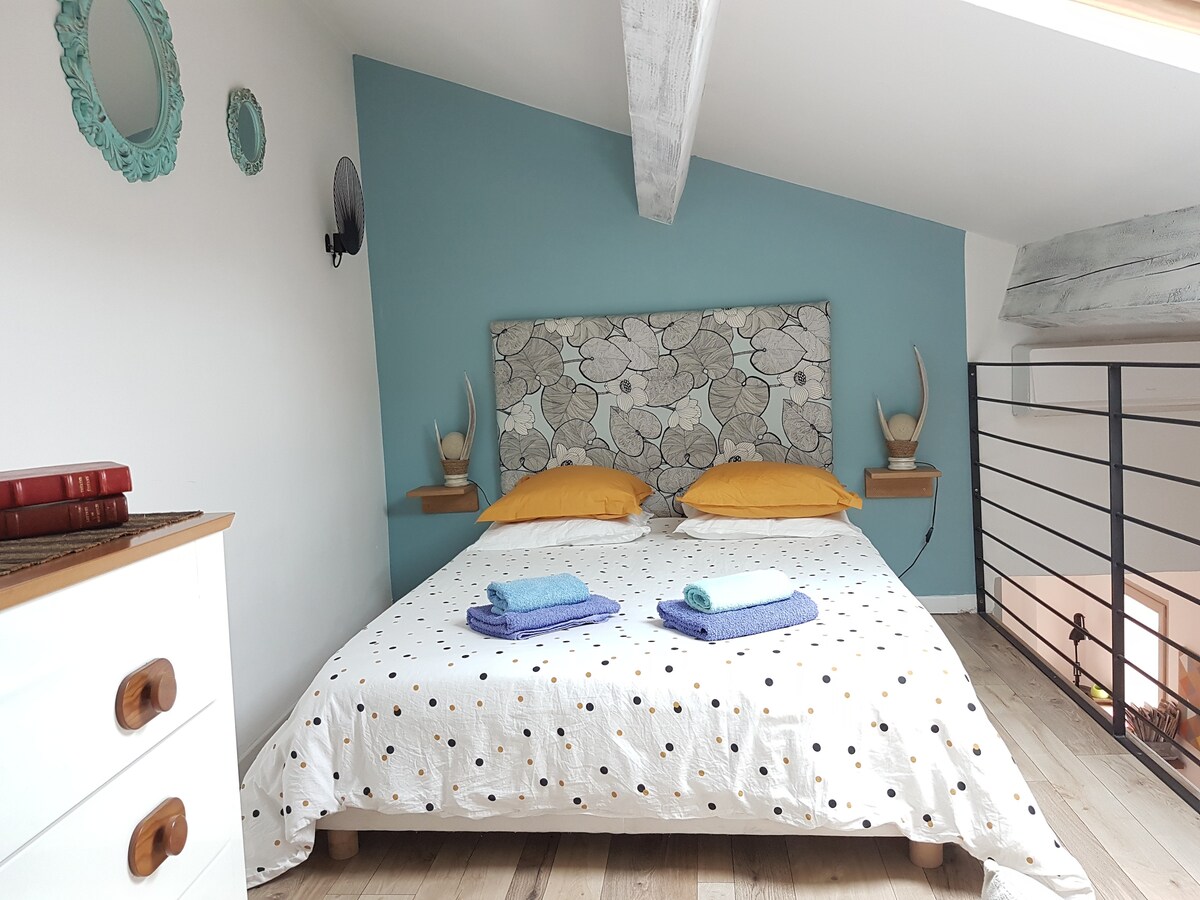 A cozy mezzanine sleeping area features a spacious bed with a patterned headboard and yellow accent pillows. Soft bedding is complemented by two rolled towels placed neatly at the foot of the bed. Light wooden floors and a light blue accent wall enhance the serene ambiance.