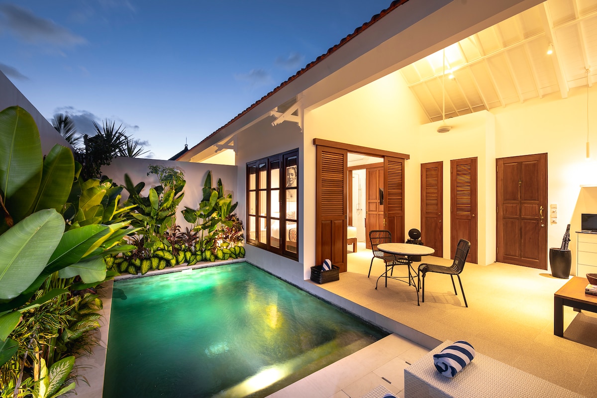 A serene private pool is bordered by tropical plants, with softly illuminated surroundings at dusk. The open-plan living area features a compact dining table and chairs, inviting relaxation. Glass doors provide a seamless transition between indoor and outdoor spaces.