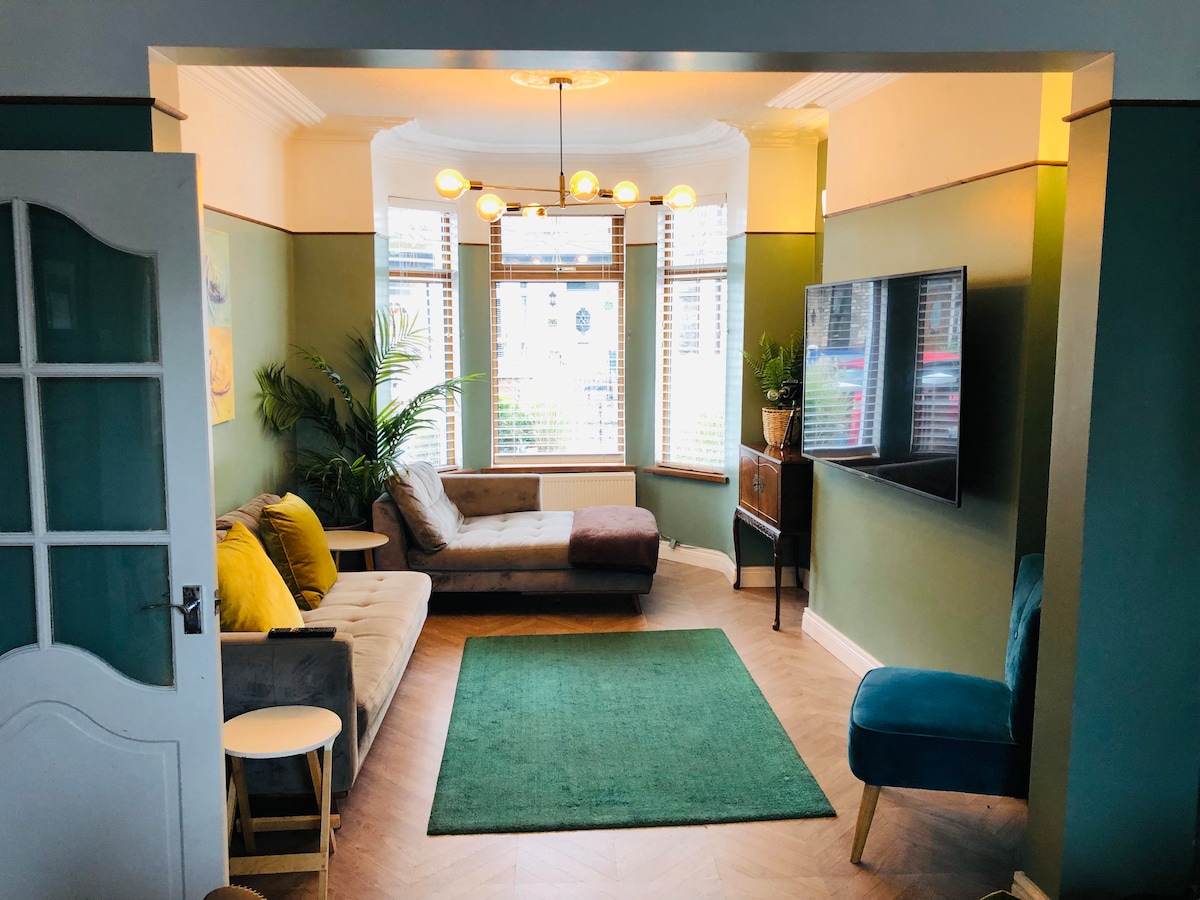 A living area features a sectional sofa and a chair, with a television mounted on the wall. Natural light enters through the large windows, creating a bright atmosphere. A green area rug sits on the wooden floor, enhancing the room's ambiance.