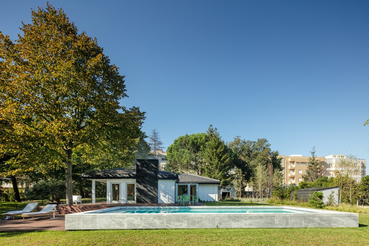 A serene outdoor area features a modern pool surrounded by well-maintained grass. The house showcases large windows and a sleek architectural design, complemented by a mature tree nearby. The setting offers a blend of natural beauty and contemporary style.